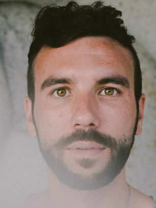A man with short dark hair and a beard stares intently. Background is a textured stone surface, creating a calm and contemplative mood.