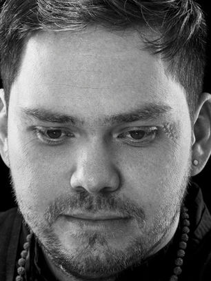 Black-and-white close-up of a serious man with an earring and beaded necklace against a dark background.