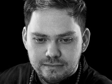 Black-and-white close-up of a serious man with an earring and beaded necklace against a dark background.
