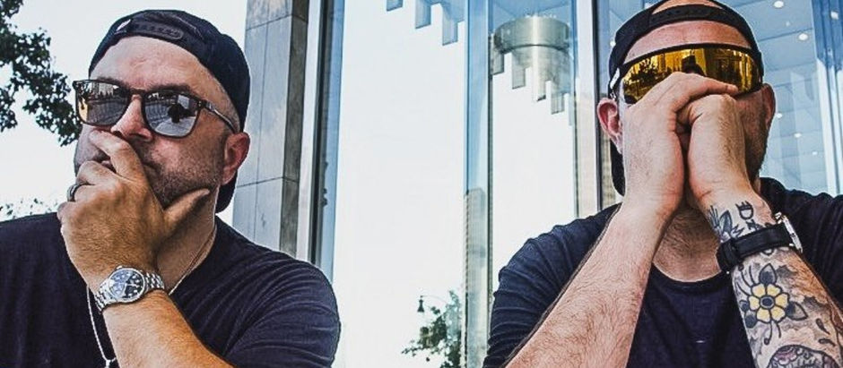 Two men in sunglasses and caps pose with hands over their mouths outside a glass building, looking thoughtful or secretive.