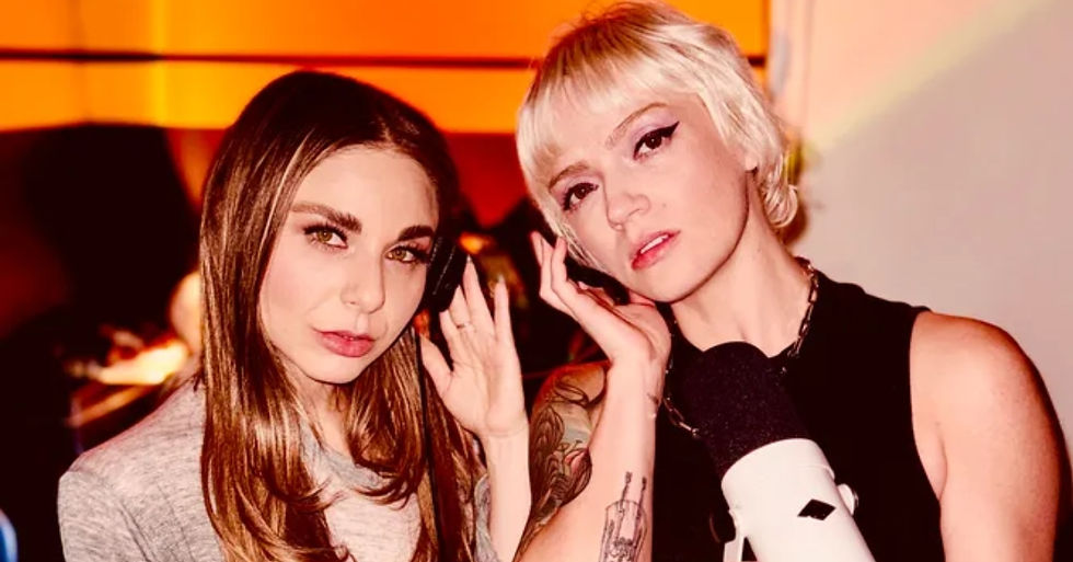 Two women with distinctive hairstyles pose with a microphone, set against a warm orange background. Mood is relaxed and engaging.
