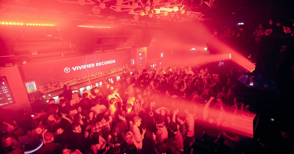 Crowded nightclub with red lighting, people dancing and socializing. "VIVIFIER RECORDS" sign visible above bar counter, lively atmosphere.
