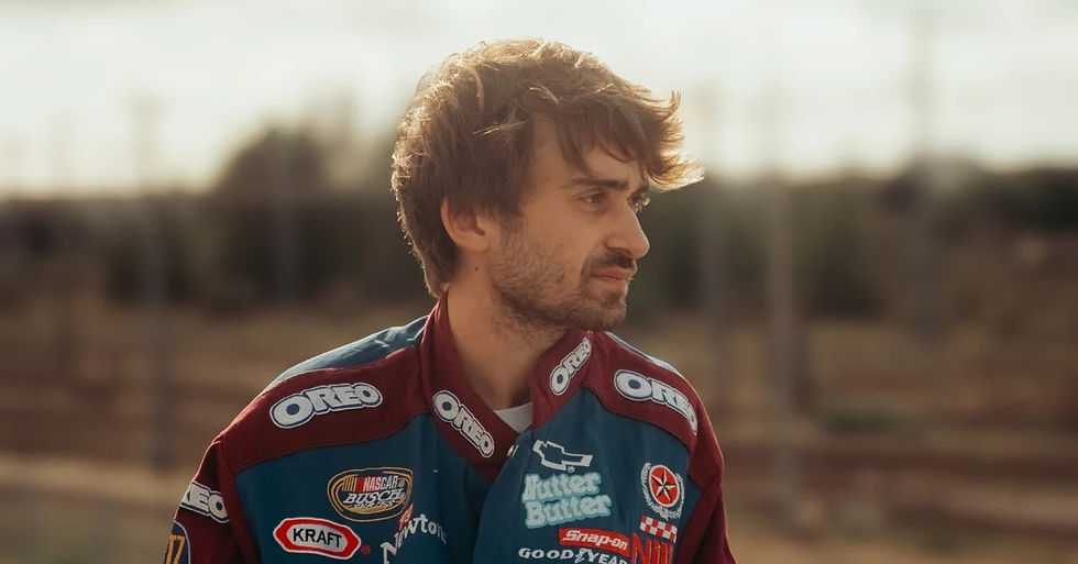 Man in a racing jacket with various sponsor logos like Oreo and Kraft, standing outdoors with a contemplative expression. Background is blurred.