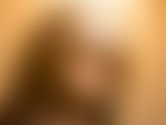 Person with long dark hair and wearing sunglasses looks confidently ahead. Warm orange lighting creates a dramatic, stylish atmosphere.