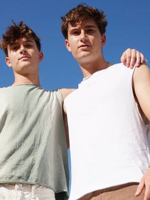 Two young men in sleeveless tops stand close under a clear blue sky, with one resting his arm on the other's shoulder, conveying friendship.