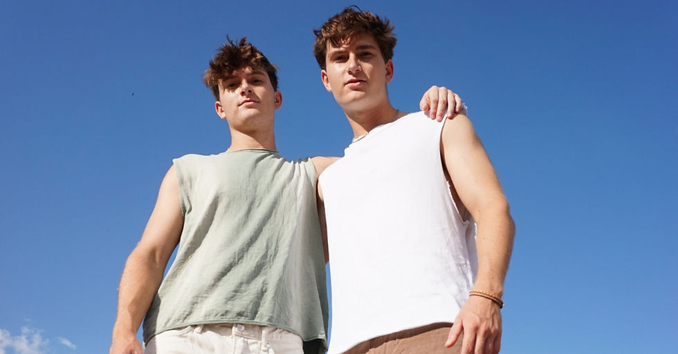 Two young men in sleeveless tops stand close under a clear blue sky, with one resting his arm on the other's shoulder, conveying friendship.