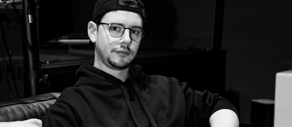 Man in glasses and cap, wearing a hoodie, sits on a sofa in dimly lit room, looking thoughtful. Black-and-white color scheme.