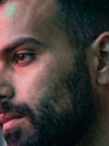 Man with beard looks thoughtful in neon-lit city setting. Soft pink and blue light reflects on his face, creating a contemplative mood.