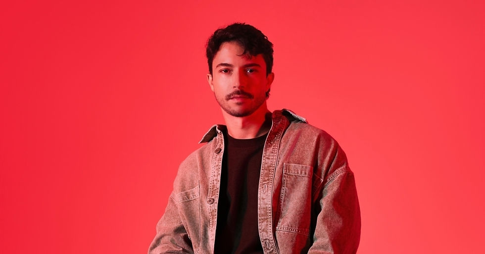 Young man in a denim shirt over black top poses against a vivid red background with a neutral expression.