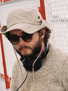 Man in sunglasses and beige hat sits on a boat deck, wearing headphones. Red railing and white mesh fence in the background. Calm expression.