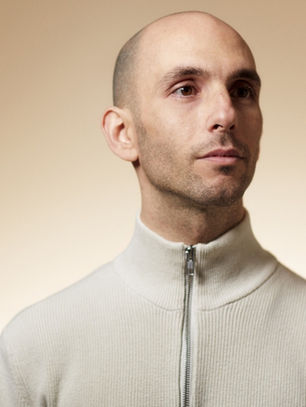 Bald man in a light zip-up top looks thoughtful against a beige gradient background. His expression is neutral and focused.