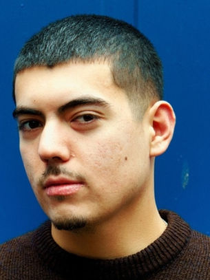 Young man with short hair and a neutral expression against a bright blue wall. Wears a brown sweater. Mood is calm and contemplative.