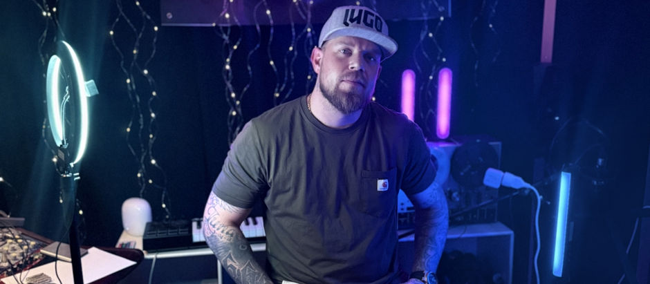 Man in a cap sits in a dimly lit studio with glowing lights and equipment. Background features fairy lights and purple accents. Serious mood.