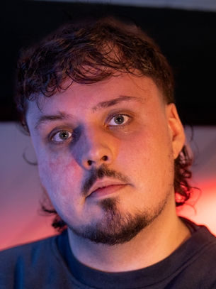 Man with a contemplative expression, standing indoors against a blurred background with blue and red lighting. He has short, curly hair and a goatee.