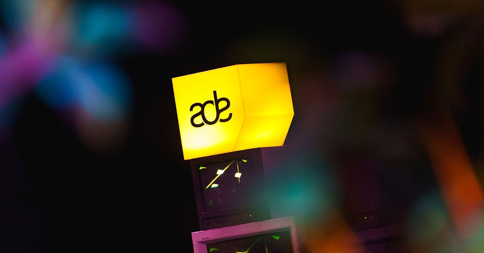 Bright yellow cube with "ade" text, set atop stacked monitors. Neon colors blurred in the background, creating a vibrant atmosphere.