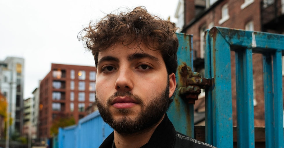 Close-up of a young bearded man with curly hair standing by a blue metal gate in a city, looking calmly at the camera.