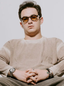 Man in beige sweater and sunglasses sits with folded hands, neutral expression. Gray background, casual setting, wearing a watch and bracelet.