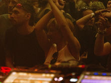 People dancing energetically at a dimly lit party, arms raised. A DJ booth is visible in the foreground. Mood is lively and vibrant.