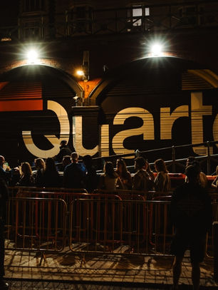 Crowd lined up outside a venue with "Quarters" on the facade. Illuminated by streetlights, the scene has a lively night-time vibe.