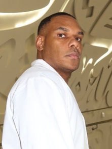 Man in white shirt stands in a patterned room with abstract designs on walls, looking serious. Beige tones dominate the background.