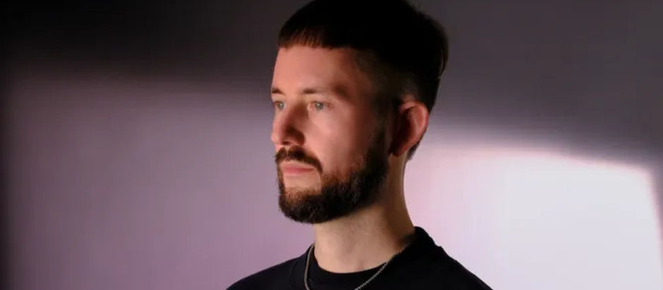 Bearded man in a black shirt poses in soft purple studio light, looking off to the side with a calm, thoughtful expression.