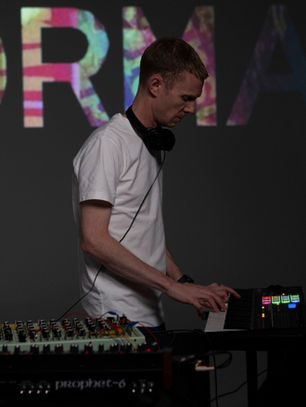 Musician in a white shirt plays keyboard with colorful lights. "KORMAC" is lit up on a dark background. Mixing equipment visible.