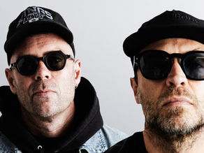 Two men in black caps and sunglasses pose against a plain gray background, one cap reading Kings; serious mood.