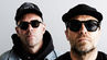 Two men in black caps and sunglasses pose against a plain gray background, one cap reading Kings; serious mood.