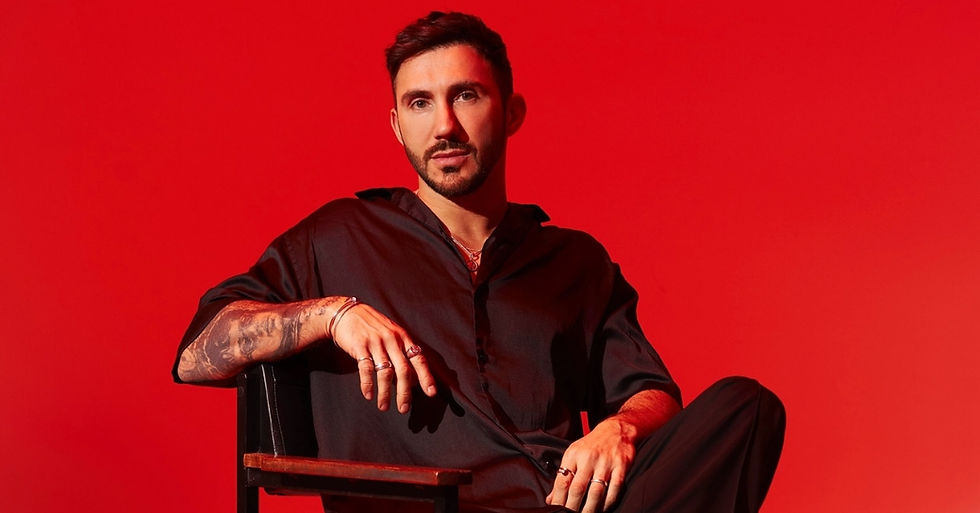 Man with tattooed arm sits in a chair against a vivid red background, wearing a black shirt and looking serious.