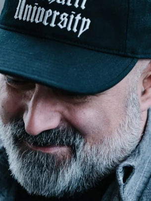 Close up portrait of a bearded man looking down while wearing a black cap and grey shirt.