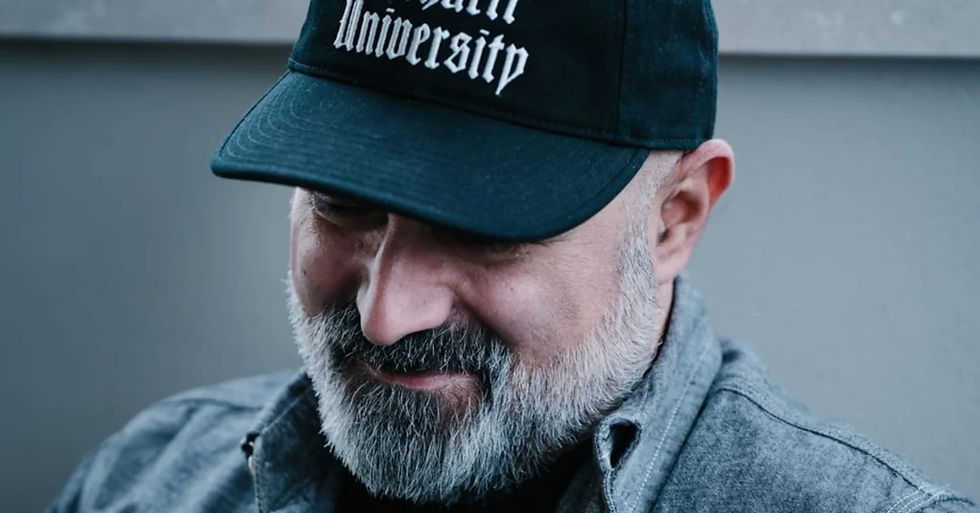 Close up portrait of a bearded man looking down while wearing a black cap and grey shirt.