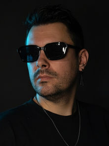 Man wearing sunglasses and black shirt with a necklace, looking to the side against a dark background. The mood is mysterious.
