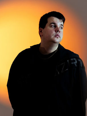 Man in a black jacket stands before a glowing orange spotlight, looking off to the right with a calm, pensive expression.
