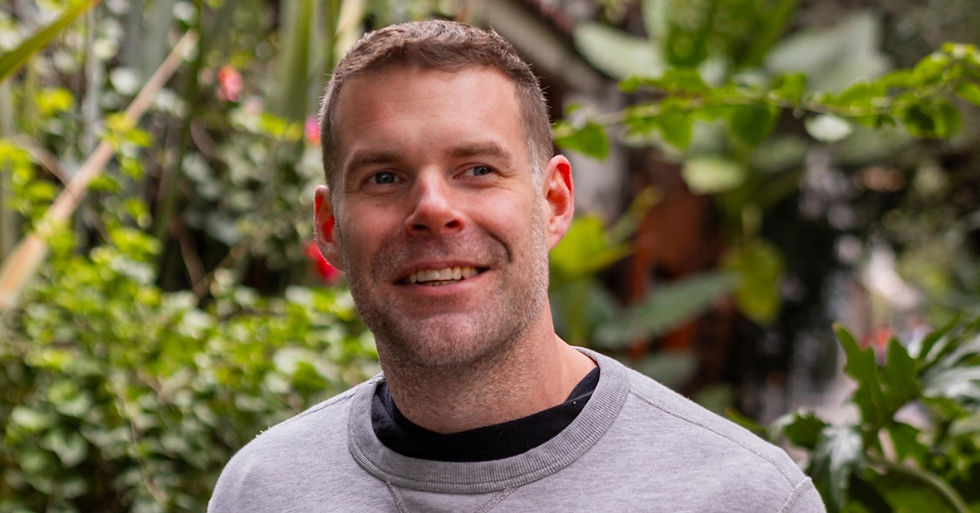 Smiling man in a lush garden or greenhouse, wearing a gray sweatshirt, with green foliage blurred behind him.
