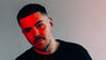 Man with short hair and stubble gazes at camera, illuminated by red light on part of face, set against a neutral gradient background.