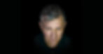 Close-up portrait of a serious middle-aged man with gray hair against a black background, lit from the front.