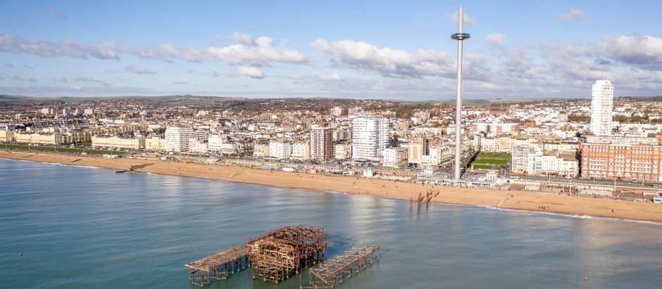 Aerial view of Brighton, showing the ruined West Pier in the calm sea, beach, and cityscape with the i360 tower under a blue sky.