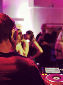 DJ wearing headphones mixes music at a lively party. People dance under vibrant purple lighting in a crowded room with energetic vibes.