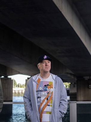 Three men stand under a large bridge by the water, looking in different directions on a cloudy day.