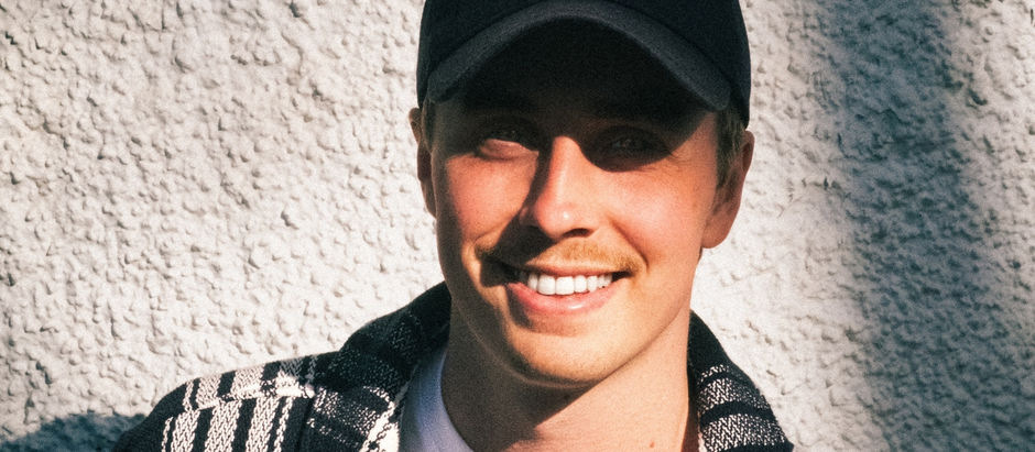 Smiling person wearing a black cap and patterned sweater stands against a textured wall in bright sunlight. Mood is cheerful and relaxed.