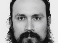 Black and white portrait of a person with long hair and a beard, looking directly at the camera. Neutral expression, plain background.