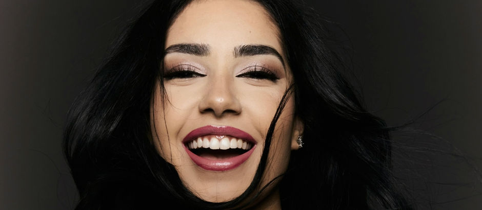 Woman with long black hair, smiling brightly against a dark background. She has bold makeup with red lipstick, exuding joy and confidence.