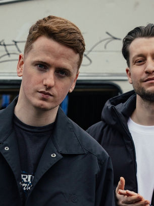 Two men in dark jackets stand before a graffitied caravan, one in front, both looking at camera with serious expressions.