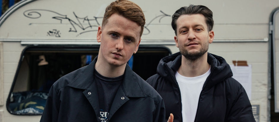 Two men in dark jackets stand before a graffitied caravan, one in front, both looking at camera with serious expressions.