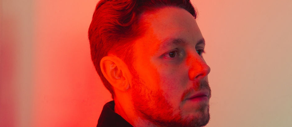 Portrait of a person with red lighting on their face, looking to the right. The background is a soft gradient of orange and red.