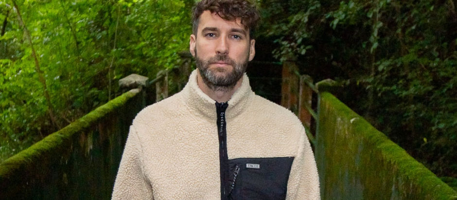 Man in a beige fleece jacket stands on a mossy bridge in a lush forest. Calm expression, surrounded by greenery. Patch reads "BYLT."