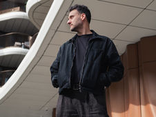 Man in black jacket with hands in pockets looks to the side, standing under a modern curved building with white and brown details.