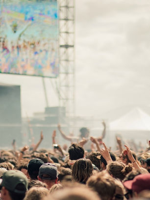 Crowd at an outdoor music festival with raised hands, stage screens, and hazy daylight under white tents.