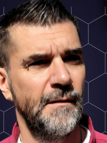 Man with a serious expression and beard, wearing a pink shirt against a black honeycomb pattern background with colorful neon accents.
