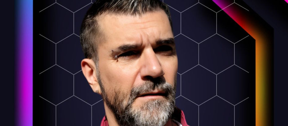 Man with a serious expression and beard, wearing a pink shirt against a black honeycomb pattern background with colorful neon accents.